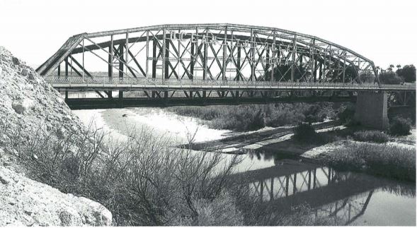 A black and white photo shows a steel truss bridge spanning a river with dry banks, surrounded by shrubs and rocky terrain, casting a distinct reflection on the water below.