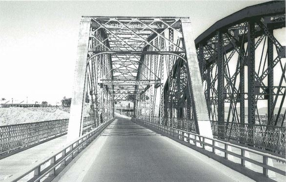 Black and white photo of an old steel truss bridge with an empty roadway, intricate lattice railings, and another similar bridge visible to the right.