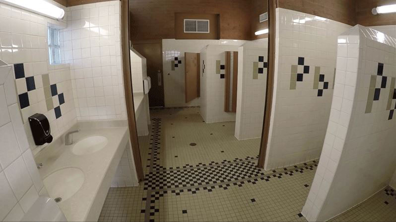 Public restroom with tiled walls and floors, two sinks with a soap dispenser, and several open stalls separated by partitions.