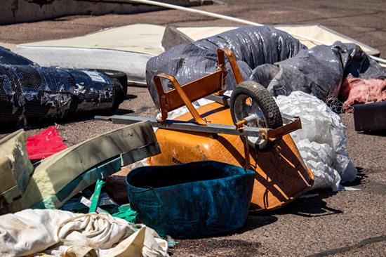 Assorted discarded items, including an overturned orange wheelbarrow, fabric, and plastic containers, scattered on a paved surface.