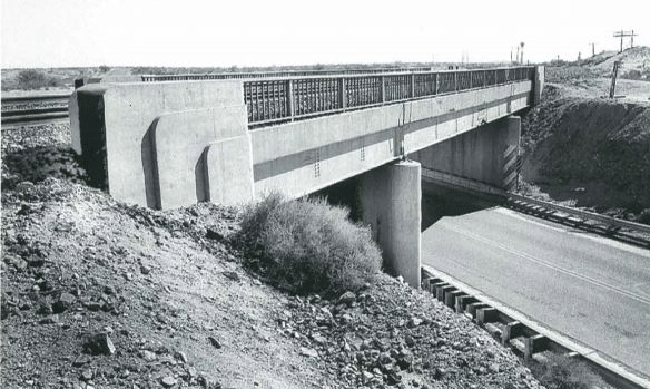 Black and white photo of a concrete bridge with metal railings crossing over a road, with rocky terrain and sparse vegetation surrounding the structure.
