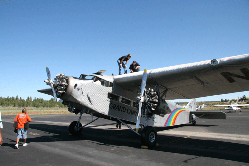 Two people work on the wing of a vintage Grand Canyon airplane parked on an airstrip under a clear sky.
