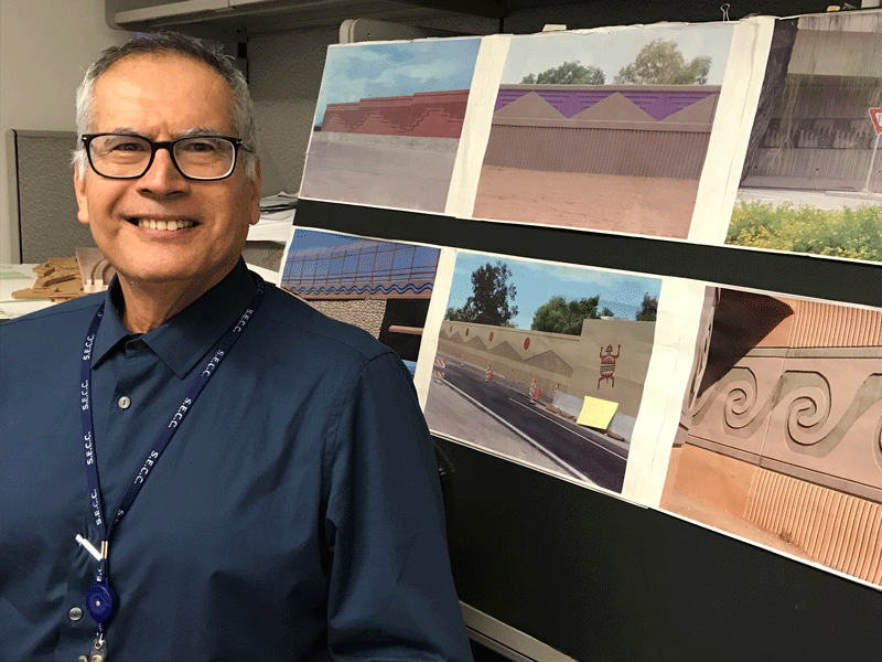 A man in glasses and a blue shirt stands next to a display of printed photos showing decorative wall designs and murals.