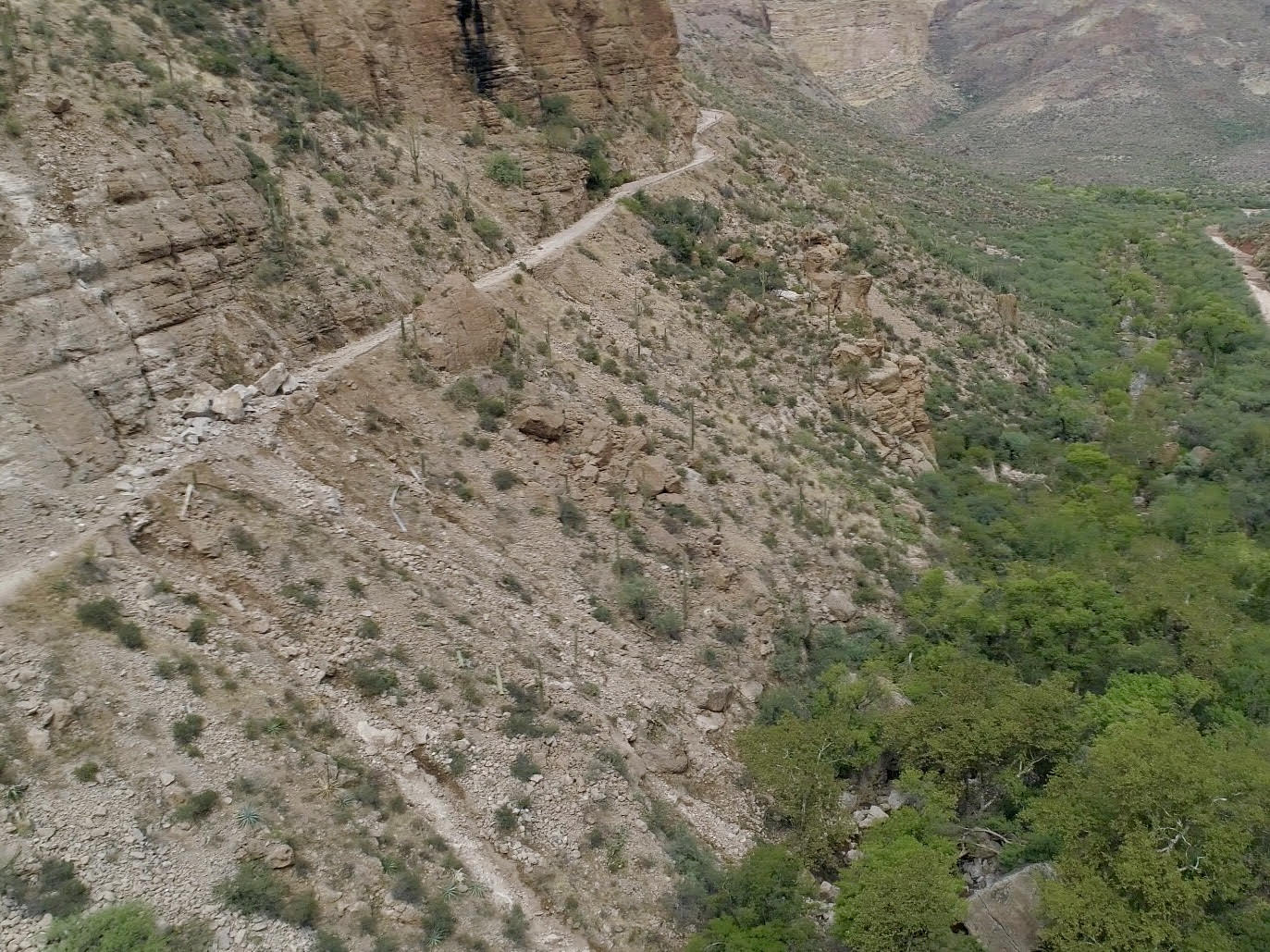 Narrow hiking trail winding along a steep, rocky canyon wall above a green valley.