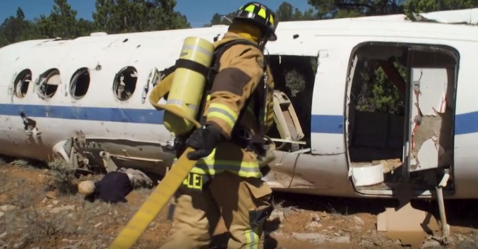 A firefighter in full gear carries a hose near the wreckage of a white airplane, with another person crouched nearby on rocky ground.