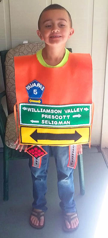 A young boy stands indoors wearing a homemade costume resembling a highway road sign with directional signs for Williamson Valley, Prescott, and Seligman.
