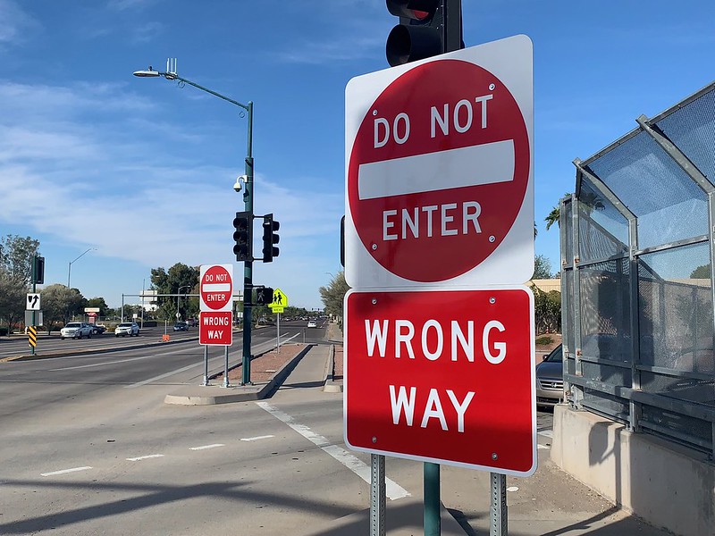 Red Do Not Enter and Wrong Way signs are posted at the entrance to a roadway, with traffic lights and a fence visible on the right side.