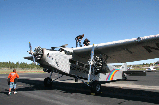 Two people stand on the wing of a silver vintage airplane labeled “Grand Canyon Airlines” with rainbow stripes, while others stand nearby on the tarmac.