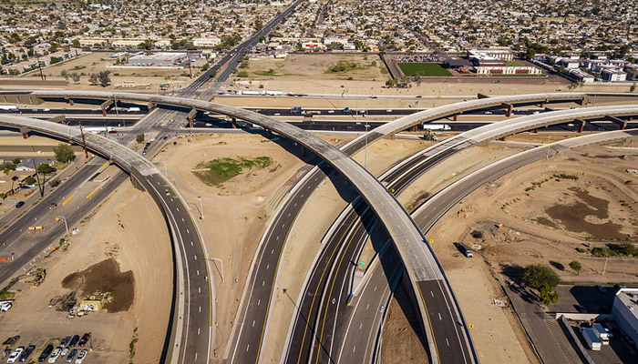 Aerial view of a large highway interchange with multiple overpasses crossing, surrounded by roads, dirt areas, and a residential neighborhood in the background.