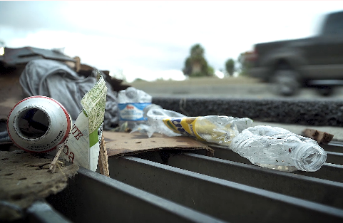 Litter including plastic bottles, a can, and cardboard are scattered on a storm drain by a roadside. A blurred truck drives past in the background.