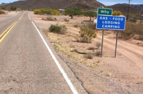 A blue roadside sign reads, Why: Gas, Food, Lodging, Camping beside a deserted highway in a dry, desert landscape with distant hills.