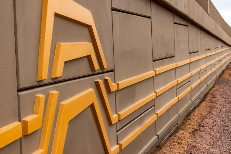 A concrete retaining wall features angular yellow geometric patterns; the wall extends into the distance with gravel at its base and a cloudy sky above.