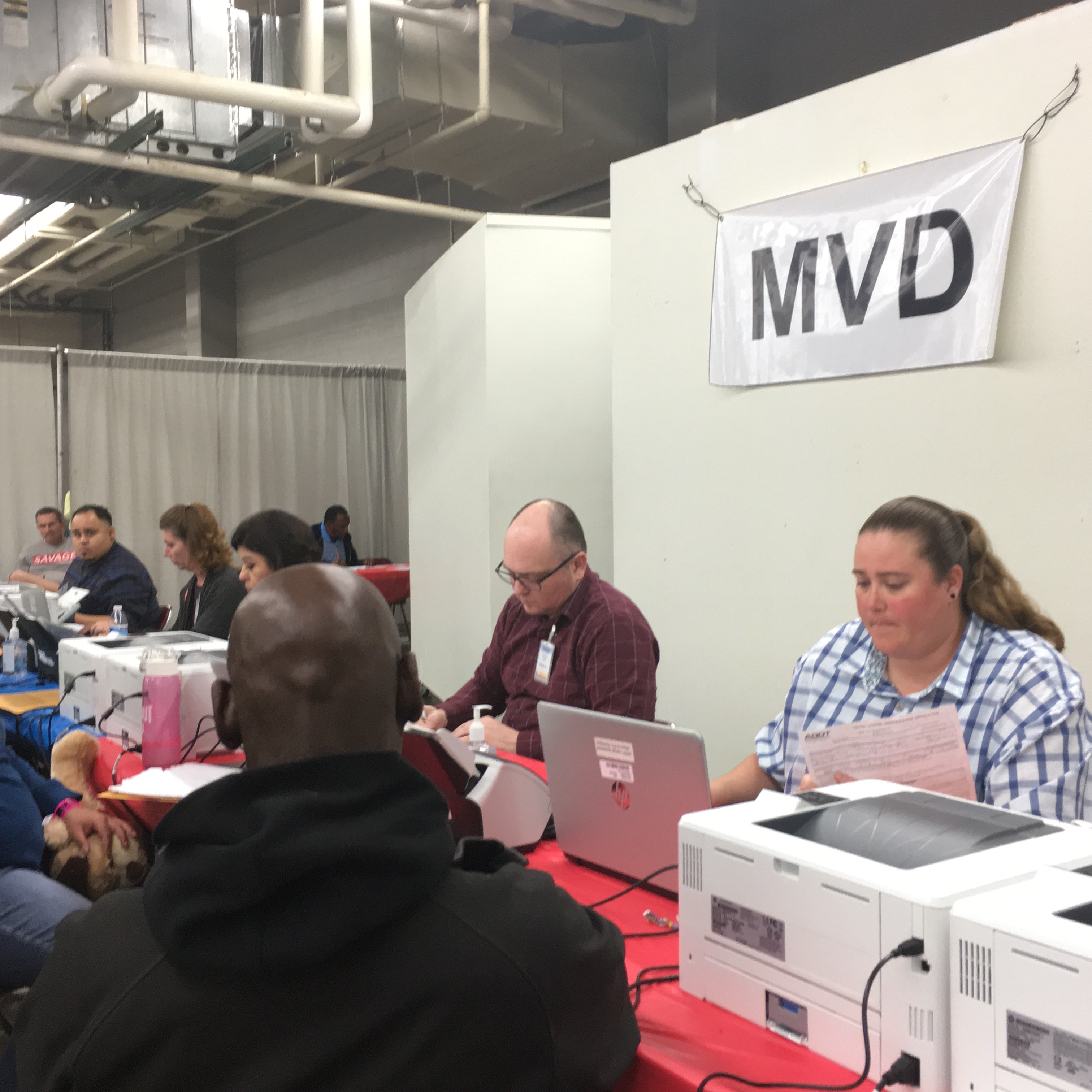 People seated at a DMV counter, staff assisting customers with paperwork and computer tasks.