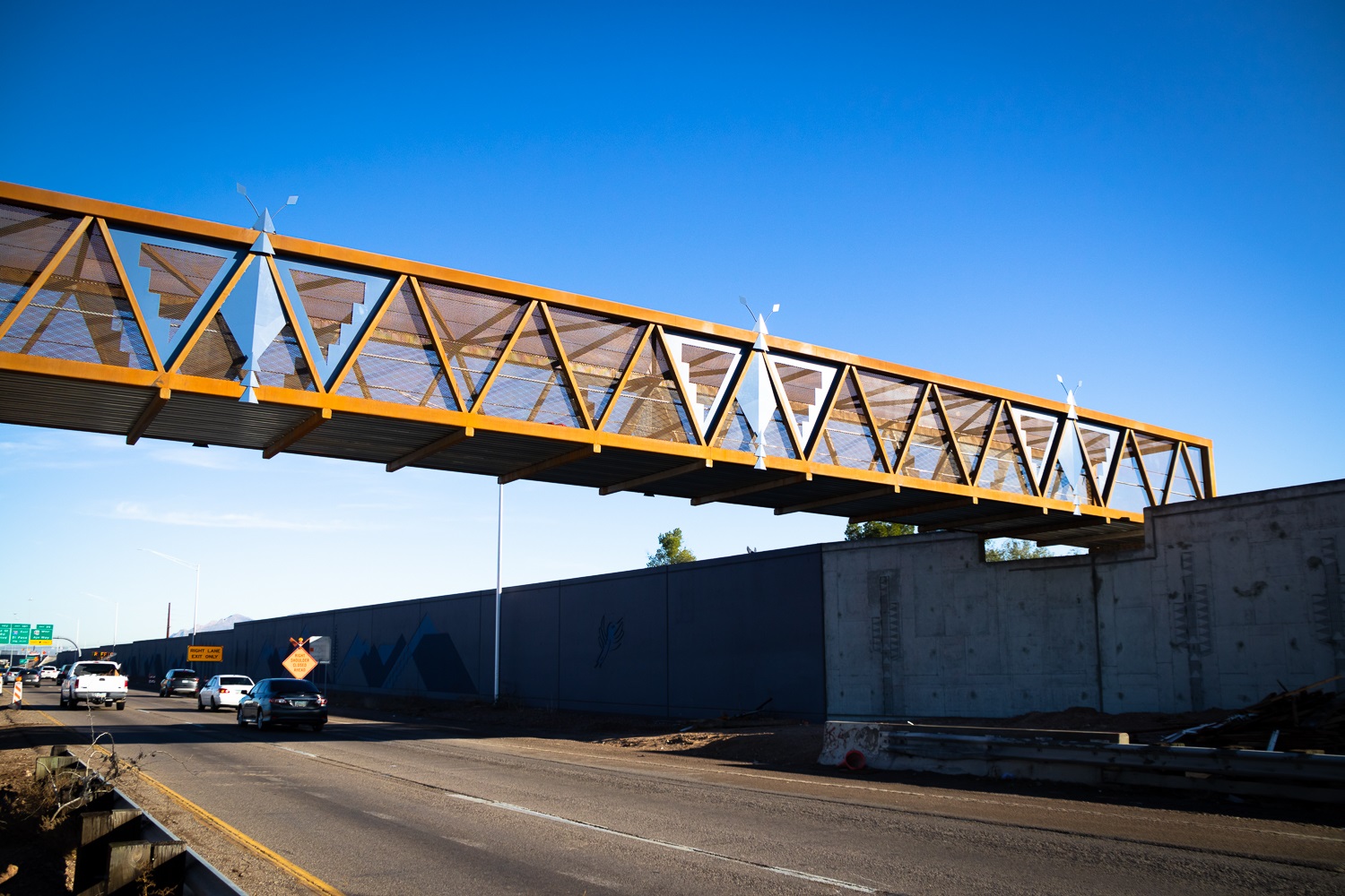 A modern pedestrian bridge with a geometric design spans over a busy highway under a clear blue sky. Cars travel beneath the bridge.