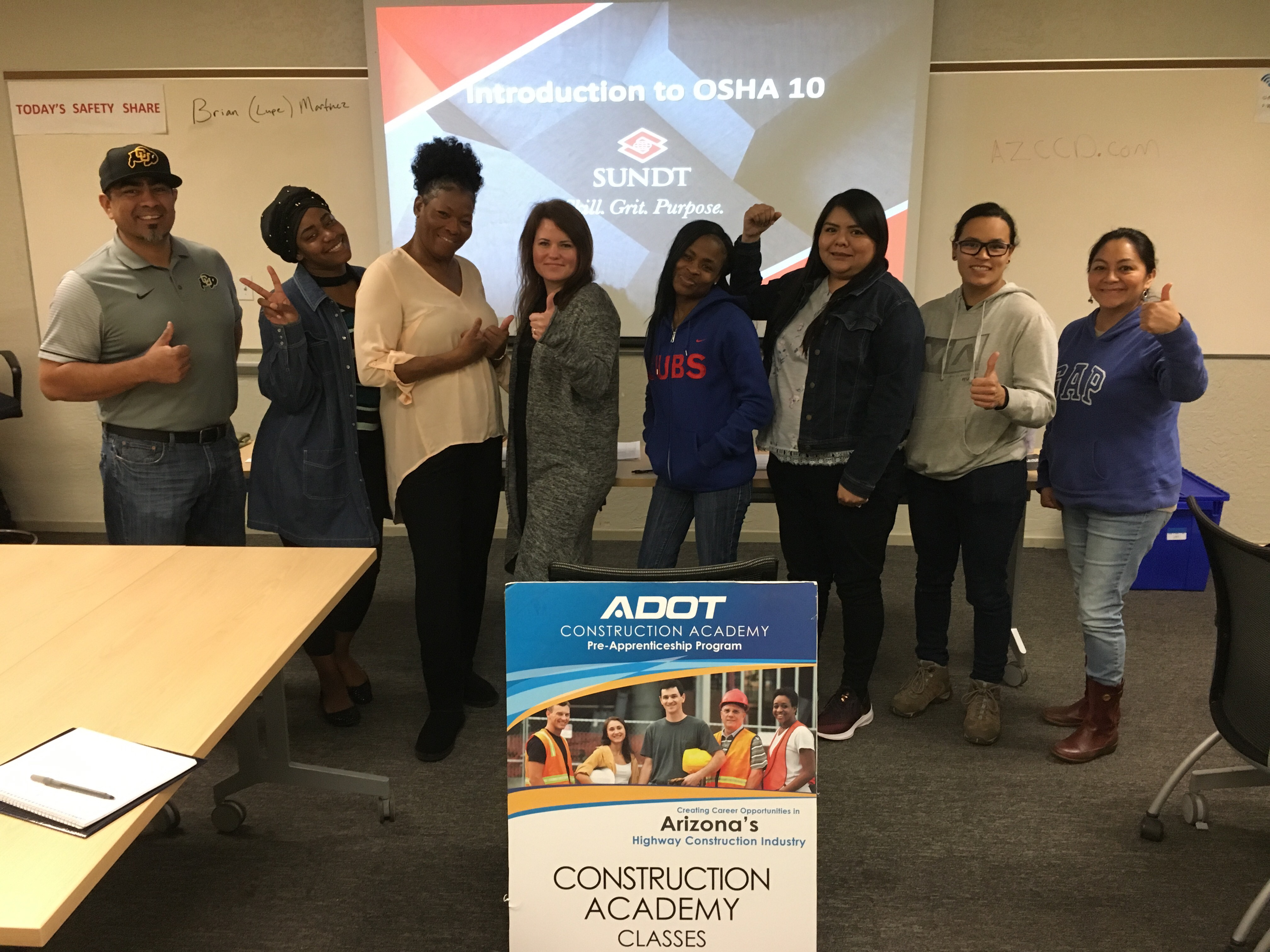 Eight people stand smiling and giving thumbs up in a classroom, with a sign for ADOT Construction Academy Classes and an OSHA 10 presentation on the screen behind them.