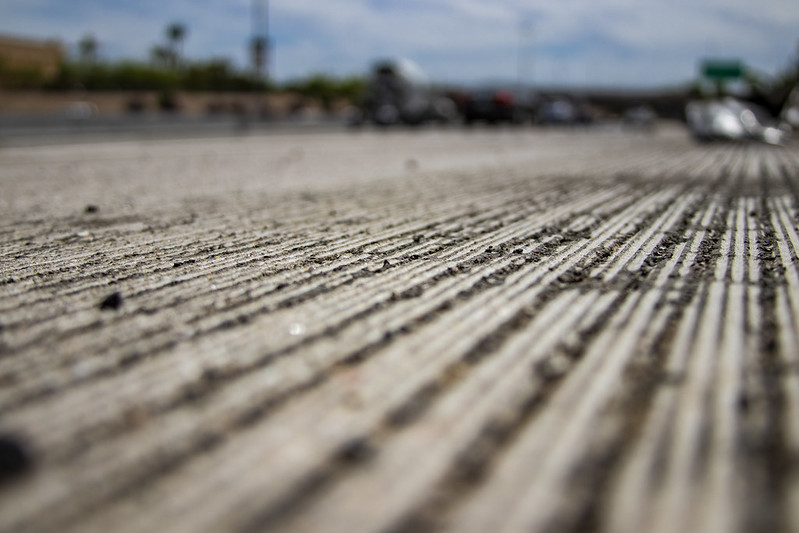 Close-up view of textured grooves on a concrete road surface, with blurred vehicles and highway scenery in the background under a partly cloudy sky.