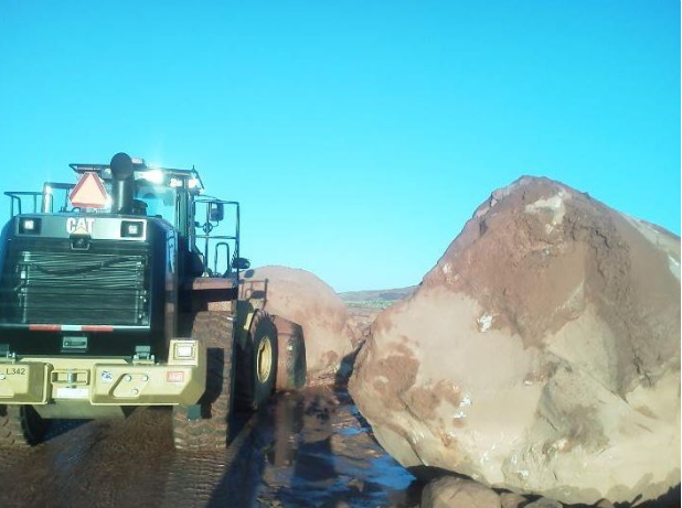A large bulldozer faces two massive boulders blocking a dirt road under a clear blue sky. The boulders appear to have fallen onto the road, obstructing passage.