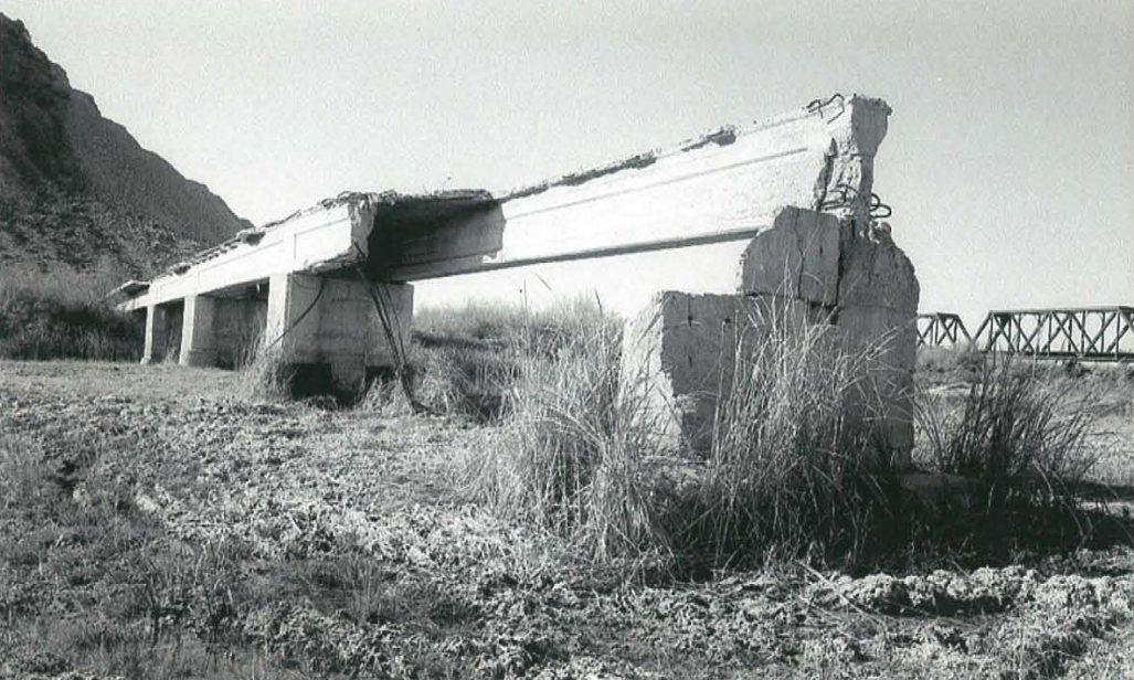 A broken concrete bridge with missing sections stands over dry, cracked ground and tall grass, with a hill and another bridge visible in the background.