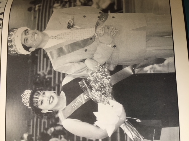 A woman and a man, both wearing sashes and crowns, stand side by side in a gymnasium. The woman holds a bouquet and both are smiling. The sashes read 1992 and King.