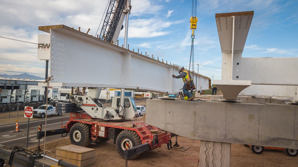 A construction worker in safety gear secures a large steel beam with bolts on a bridge structure while a crane and other equipment are visible nearby.
