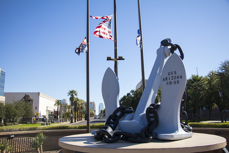 Large anchor monument with USS Arizona BB-39 inscribed, American flags flying behind, palm trees in background.