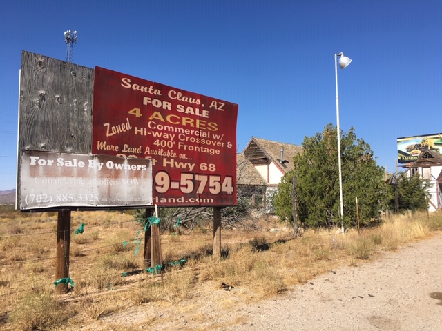Large red and white billboard advertising land for sale in Santa Claus, AZ, next to an old building and overgrown weeds on a sunny day.