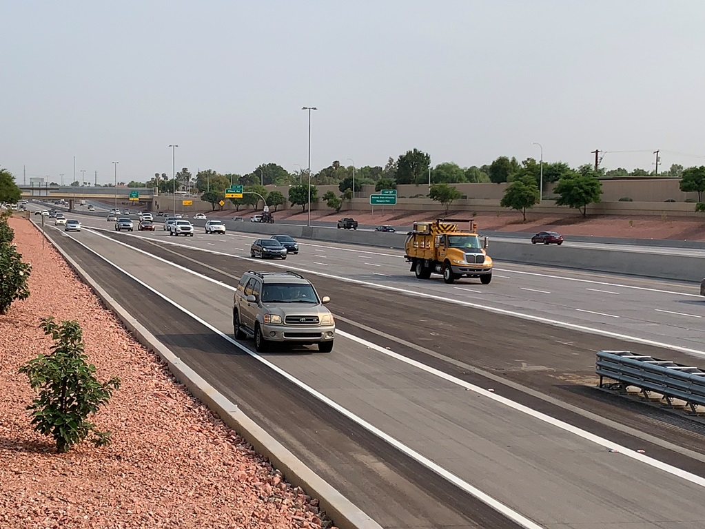 A multi-lane highway with several cars and a yellow utility truck driving under clear skies, bordered by red gravel landscaping and small trees.