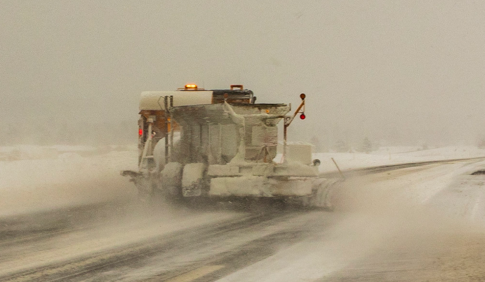 A snowplow clears a snow-covered road in low visibility during a winter storm.