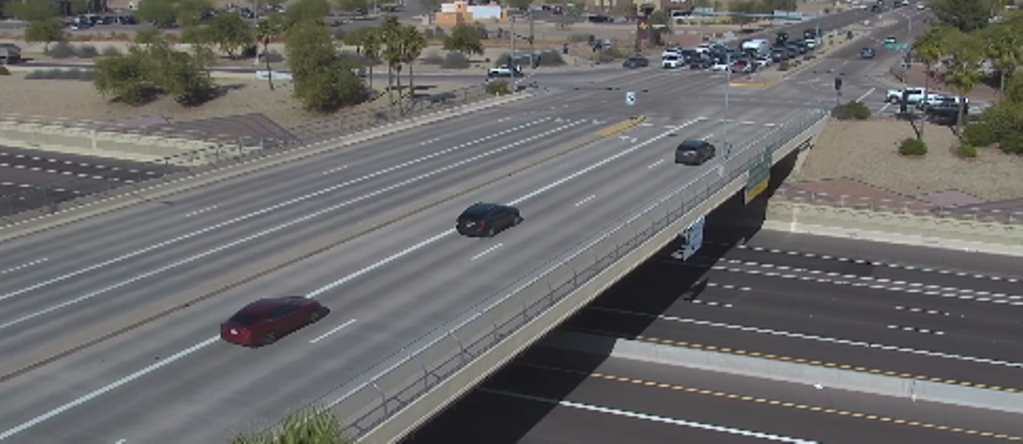 An overpass crosses above a multi-lane road with light traffic; several cars travel on both the overpass and the street below, with trees and buildings in the background.