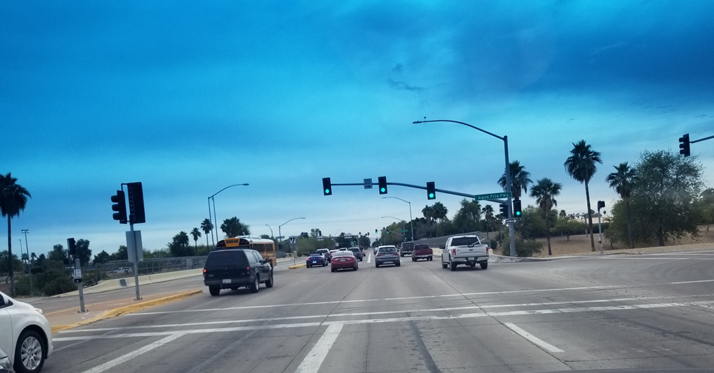 A wide intersection with several cars and a school bus driving through a green traffic light; palm trees and a cloudy blue sky are visible in the background.