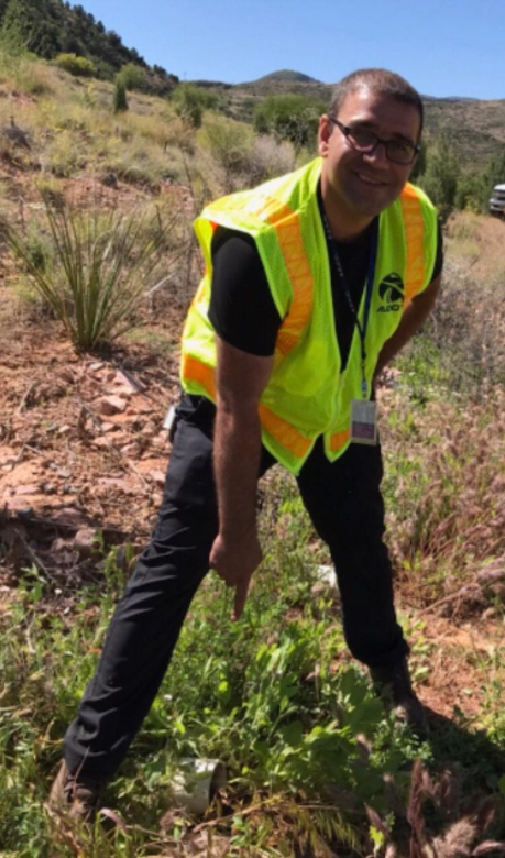 A person wearing a yellow safety vest and black pants stands outdoors on a rocky, grassy area, pointing at a patch of green plants near their feet.