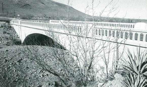 Black and white photo of a concrete bridge with arched railings spanning a dry, rocky creek bed, with sparse vegetation and a hill in the background.