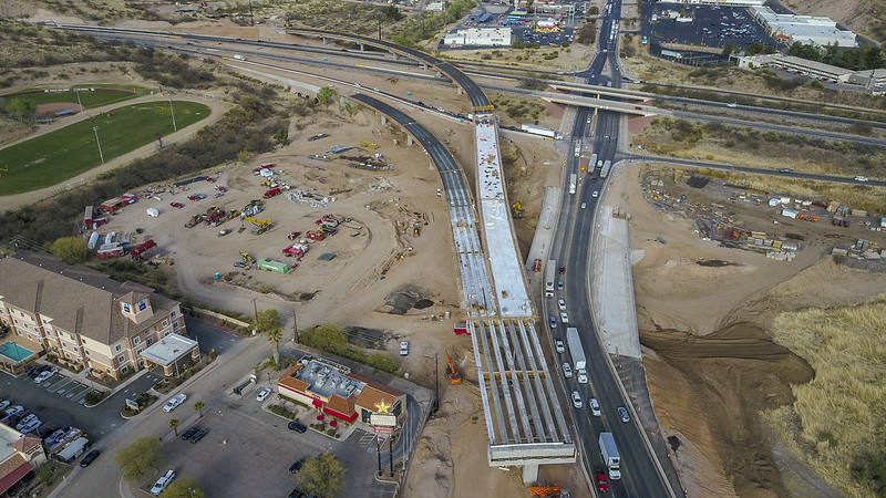 Aerial view of a highway under construction with vehicles, equipment, and buildings nearby; part of the road is unfinished while traffic moves on adjacent lanes.
