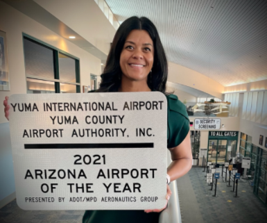 A woman stands indoors holding a sign that reads Yuma International Airport, 2021 Arizona Airport of the Year, with airport gates and security in the background.