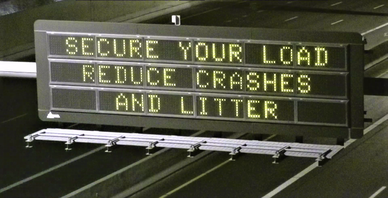 Overhead digital highway sign reads SECURE YOUR LOAD REDUCE CRASHES AND LITTER.