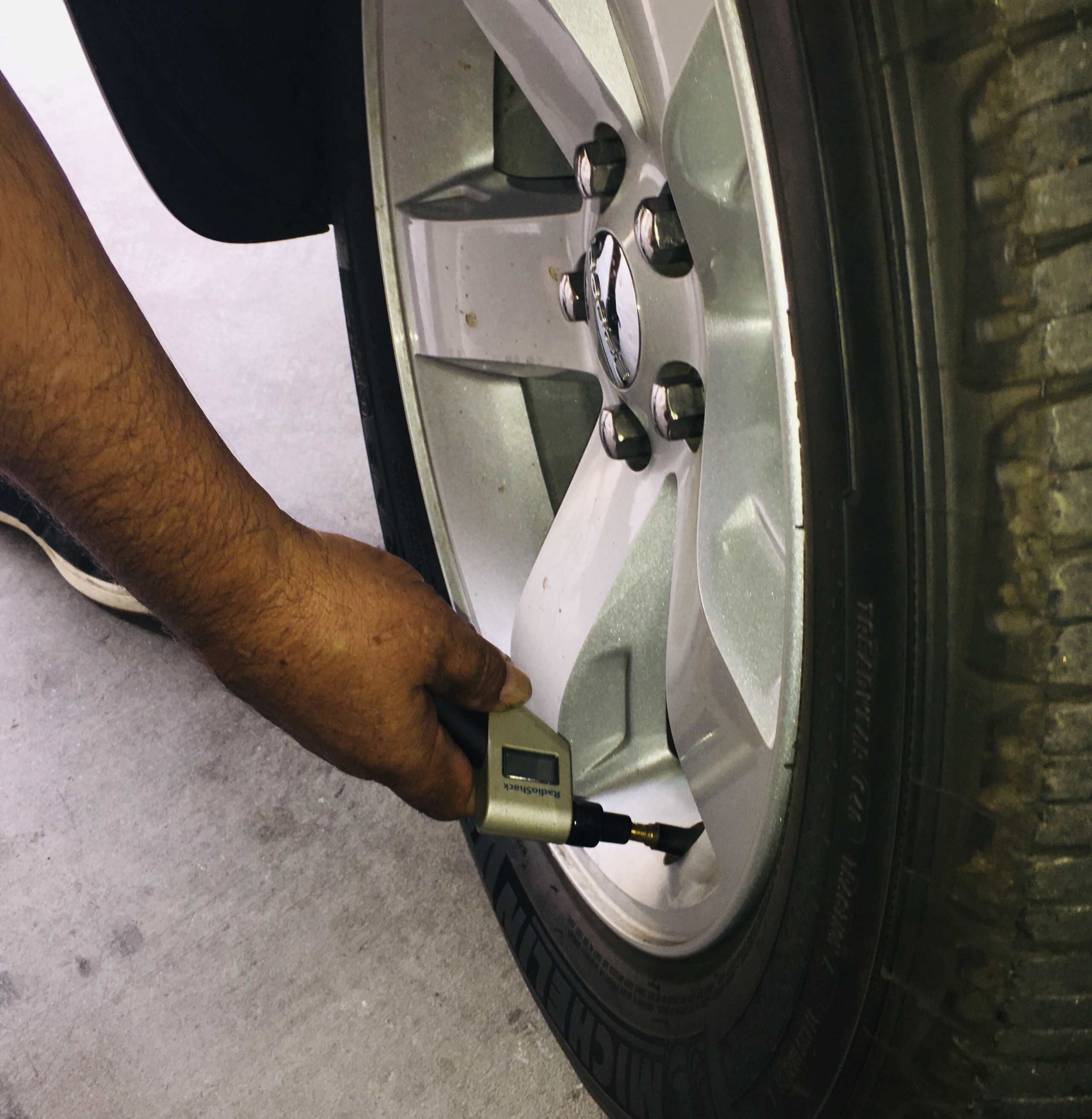 A person checks the air pressure of a car tire using a digital tire pressure gauge, with the gauge pressed against the tire’s valve stem.