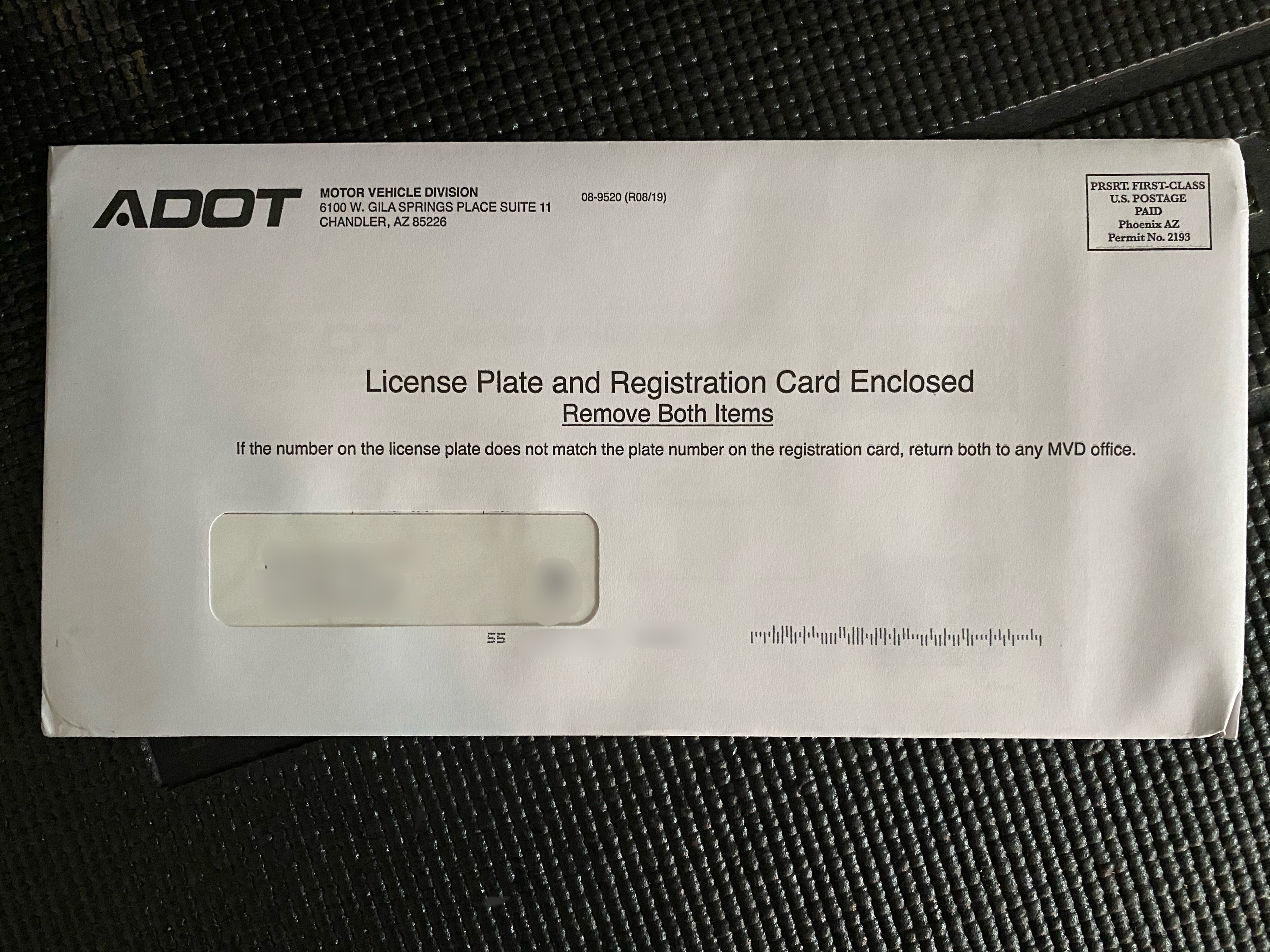 A white envelope from ADOT Motor Vehicle Division with a window showing an address, marked License Plate and Registration Card Enclosed on a black textured surface.