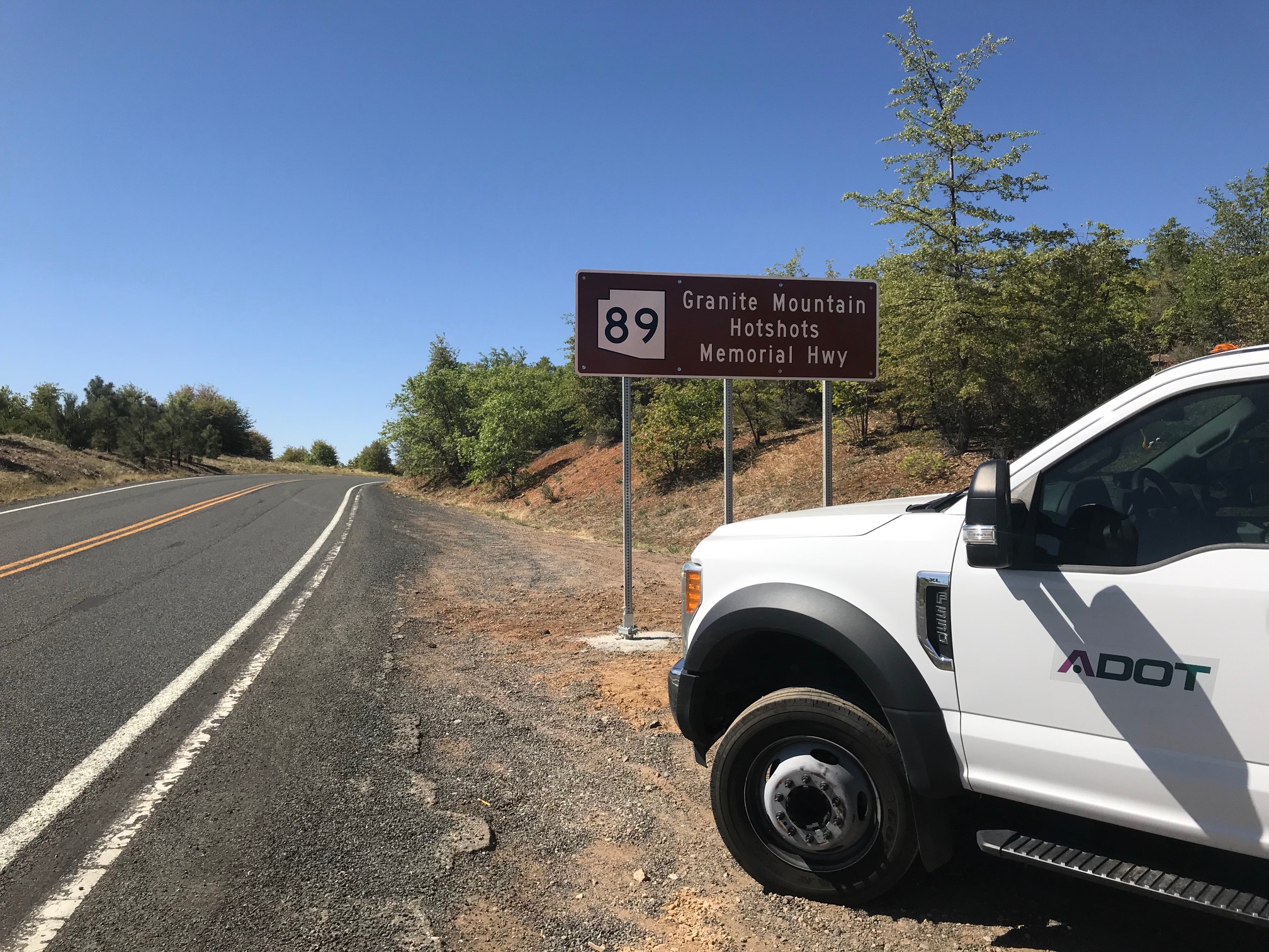 A white ADOT truck is parked on the roadside near a brown sign marking State Route 89 as Granite Mountain Hotshots Memorial Hwy, under a clear sky.