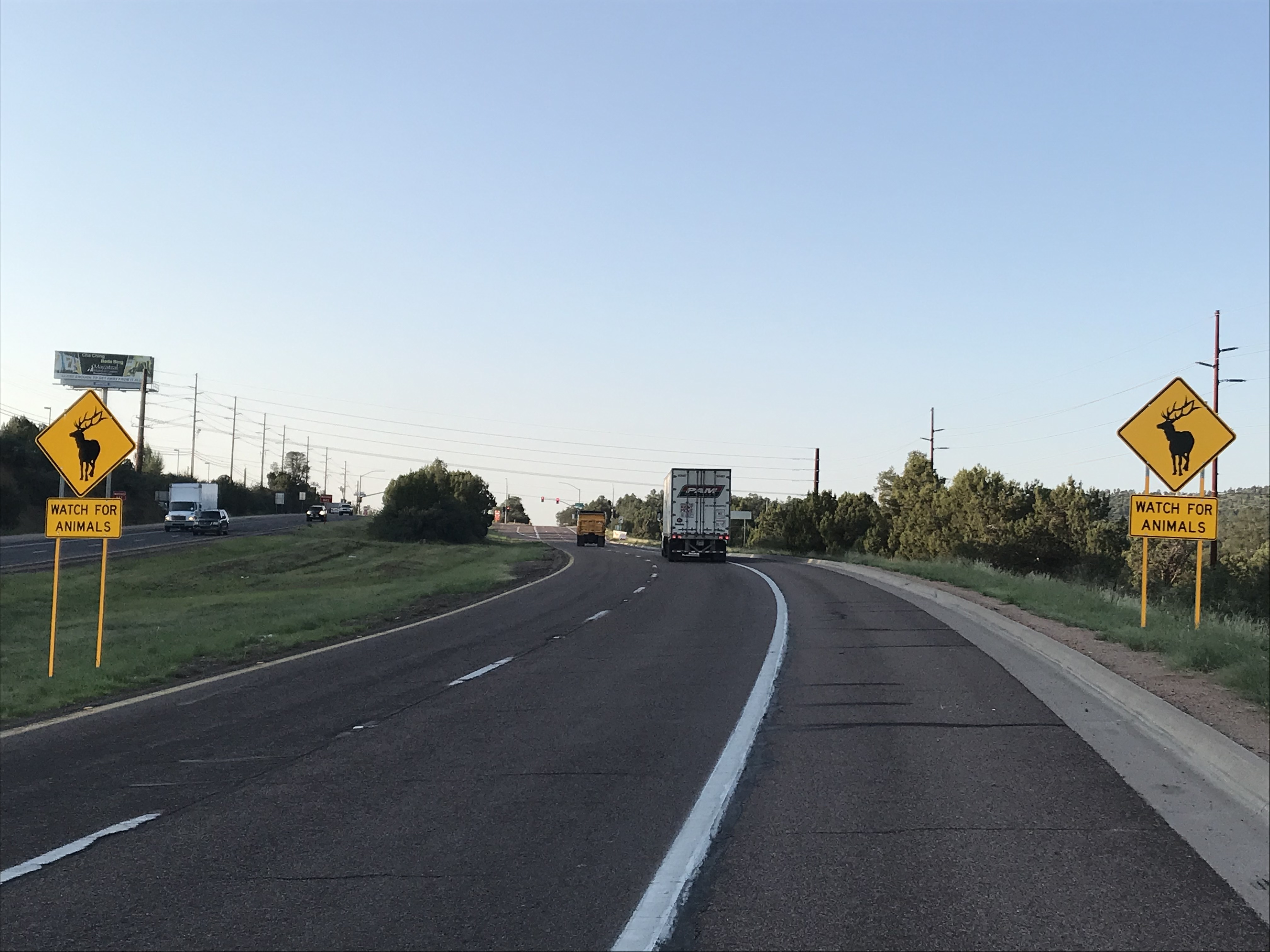 A highway with two yellow animal crossing signs reading Watch for Animals and a truck driving ahead under a clear sky. Trees line both sides of the road.