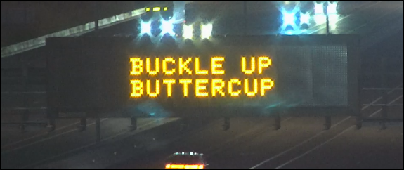 Electronic highway sign at night displays the message BUCKLE UP BUTTERCUP in bright yellow text above lanes of traffic.