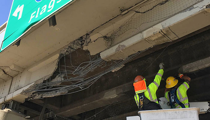 Two construction workers in safety gear inspect and repair a damaged overpass with exposed rebar and cracked concrete beneath a green highway sign.