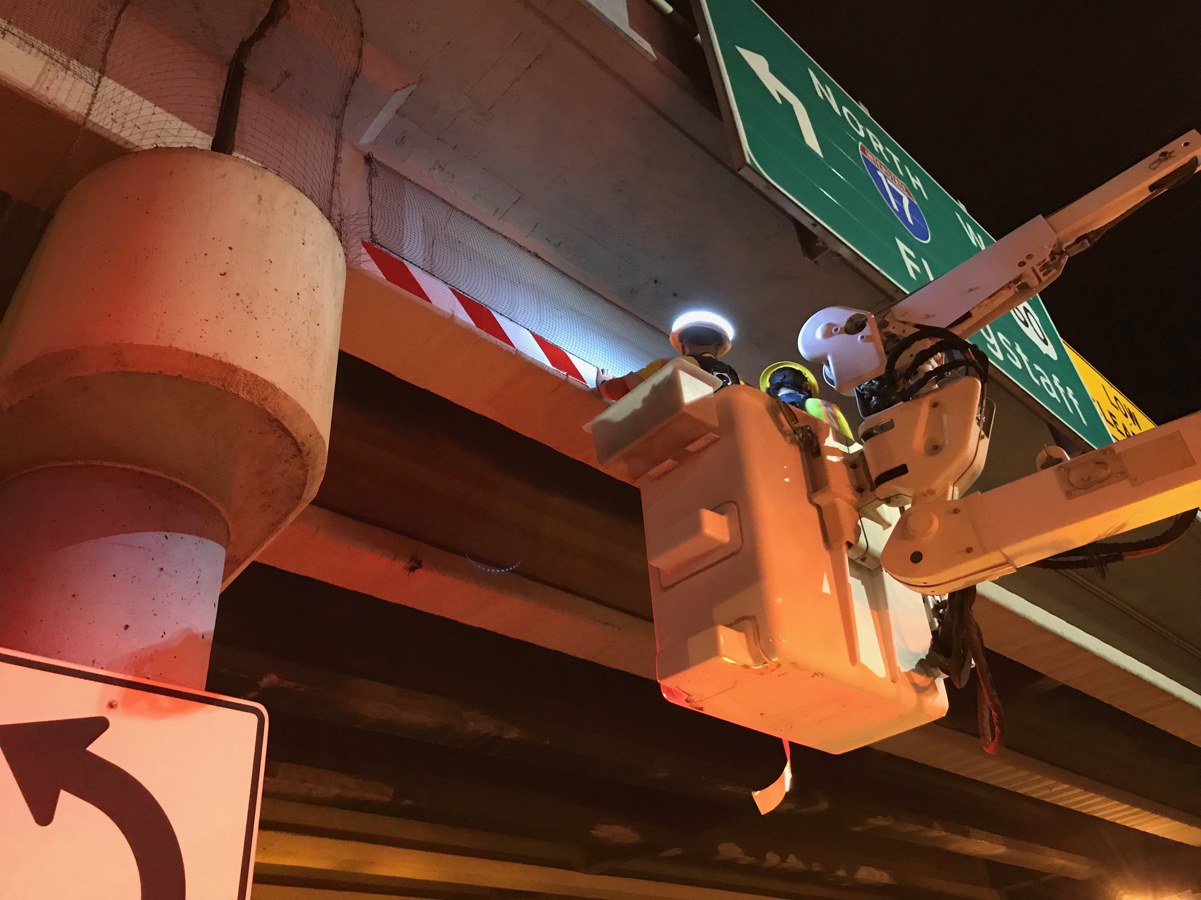 A worker in a bucket places reflective tape on the underside of a bridge deck. The scene is illuminated by work lights.