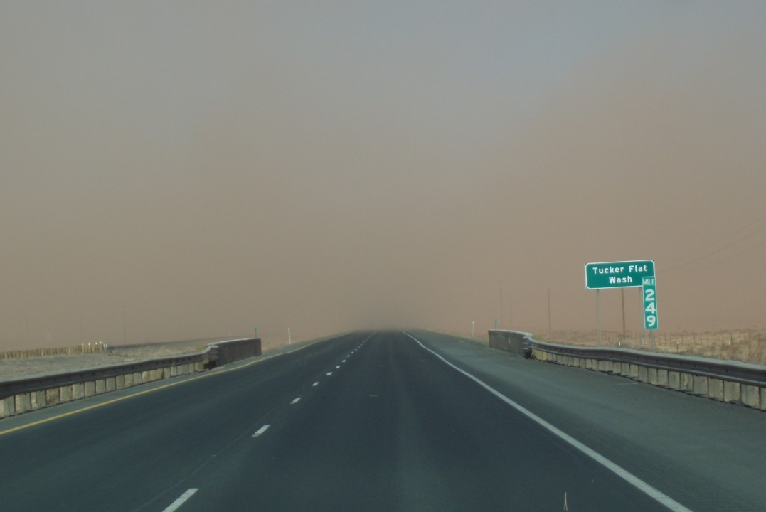 A highway stretches forward under a hazy sky with reduced visibility. A green road sign reads Tucker Flat Wash and 242.