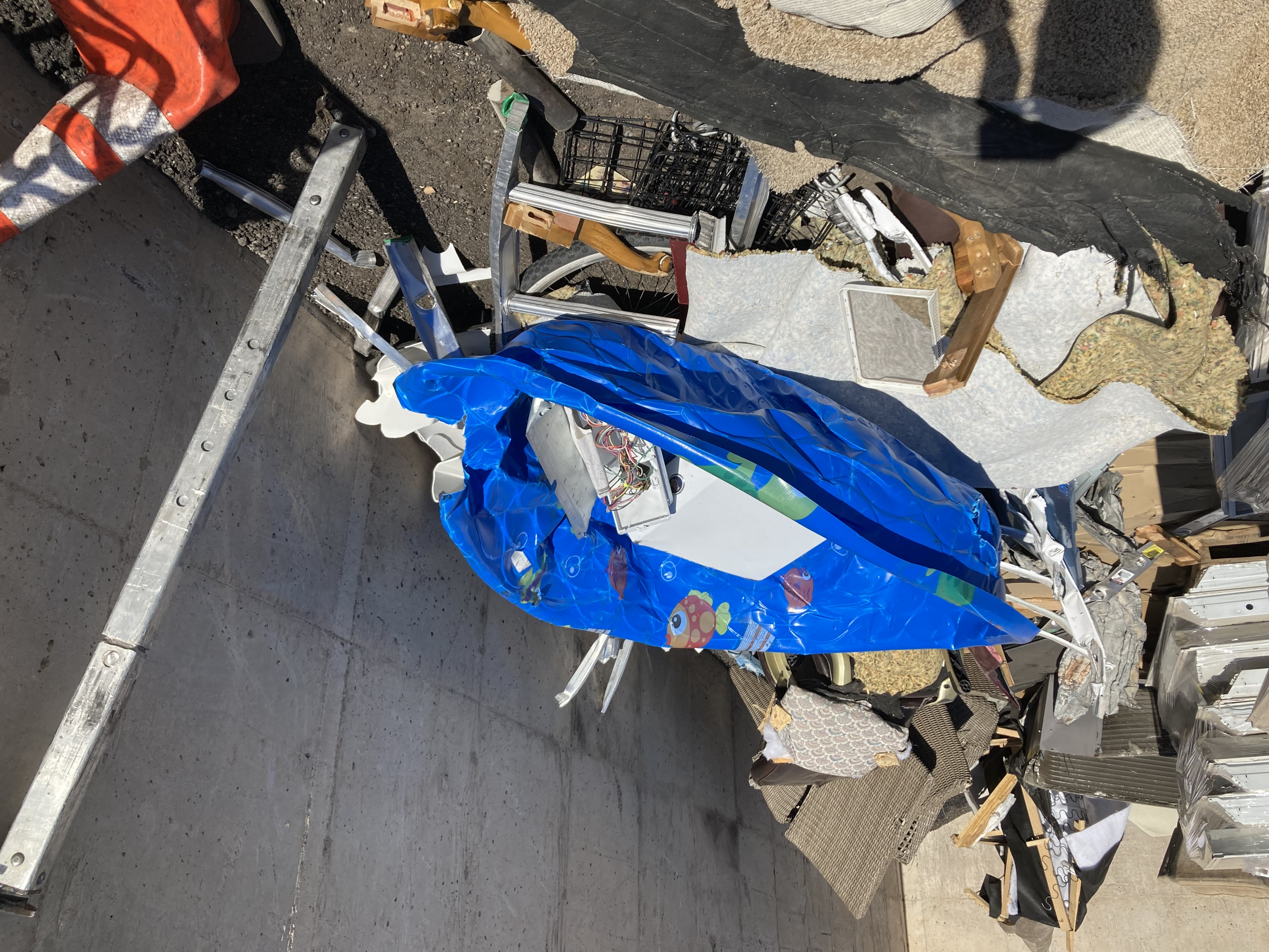 A blue inflatable kiddie pool decorated with with cartoon fish designs sits among various discarded household items in a dumpster under bright sunlight.