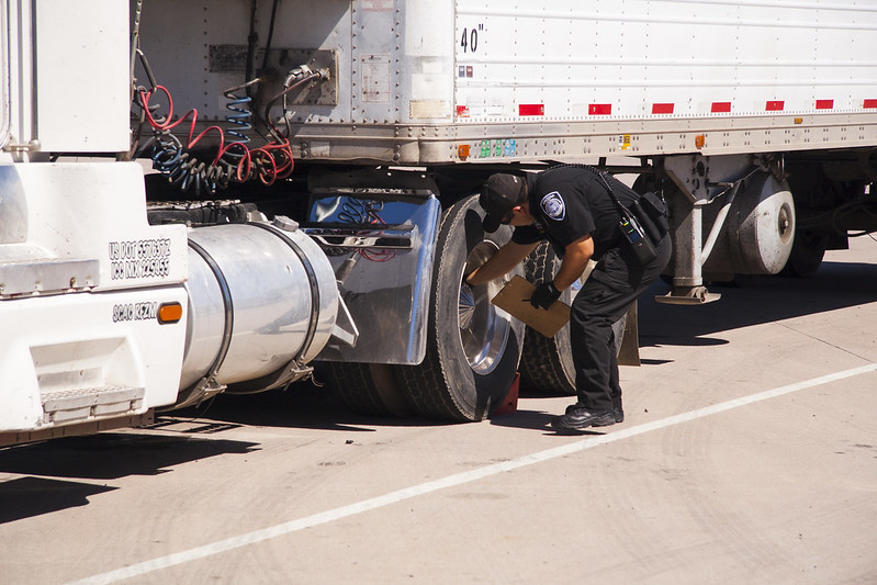 A police officer inspects the tires of a parked semi-truck on a sunny day.