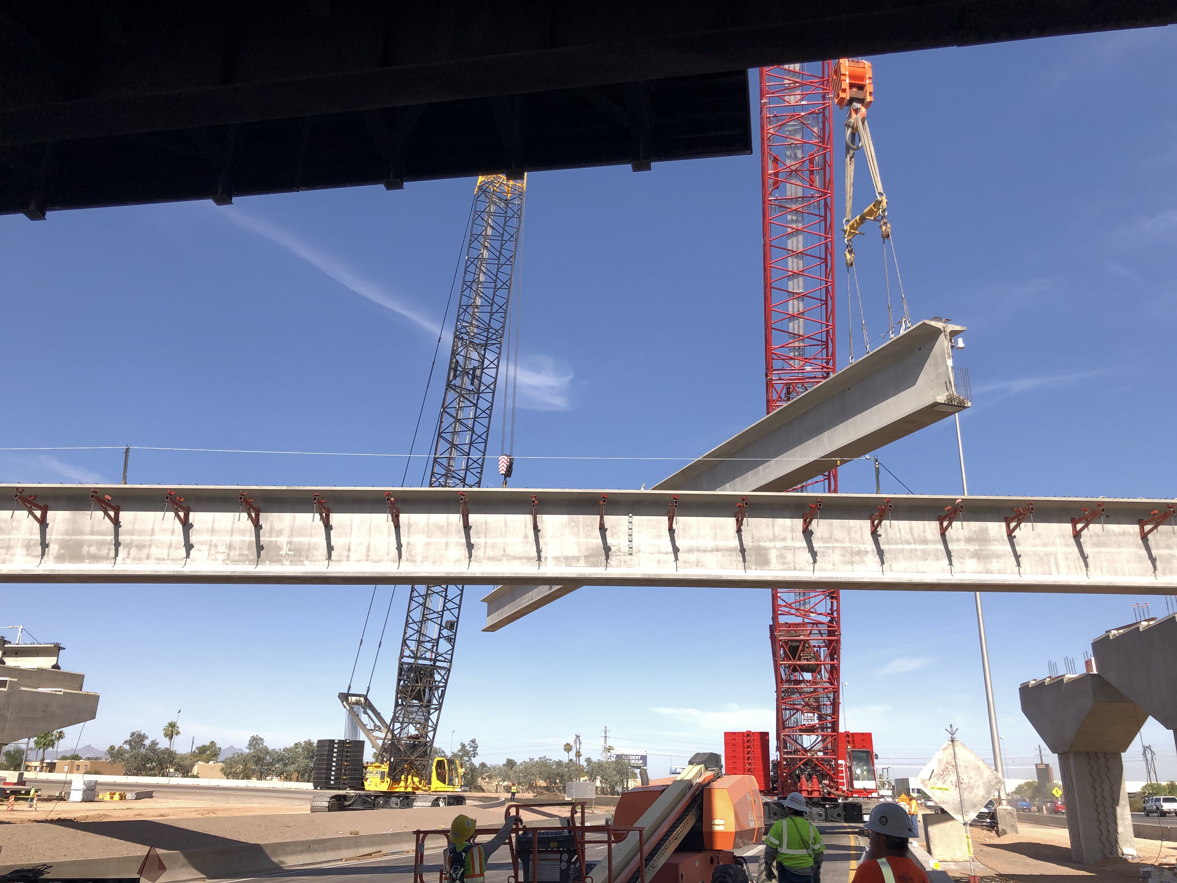 A large crane lifts a concrete bridge beam into place at a construction site, with workers in safety gear observing below.