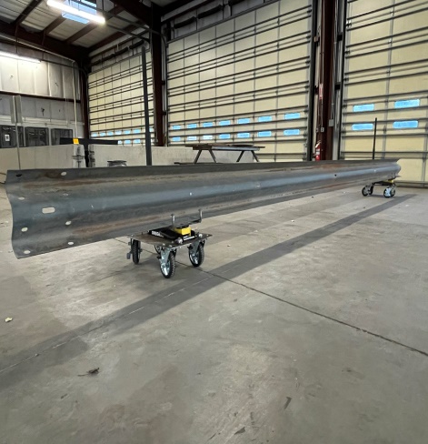 A long metal guardrail rests on two rolling carts inside a spacious industrial warehouse with large doors and concrete floors.
