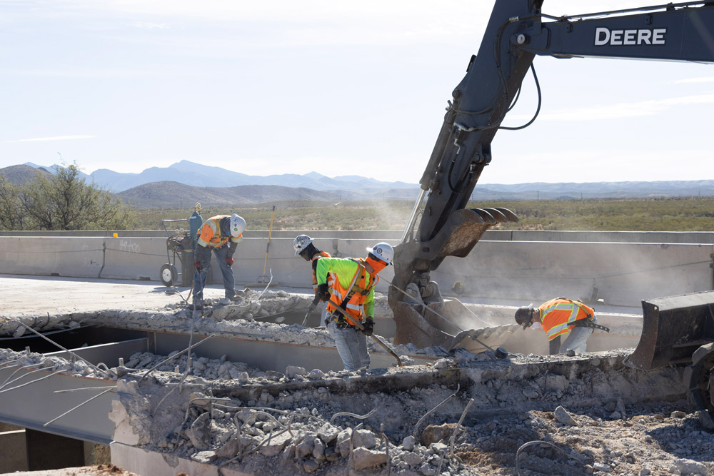 Construction crew in safety gear working beside an excavator as it breaks apart the bridge deck during the I-10: Amole TI and UPRR Bridge Rehabilitation Project.