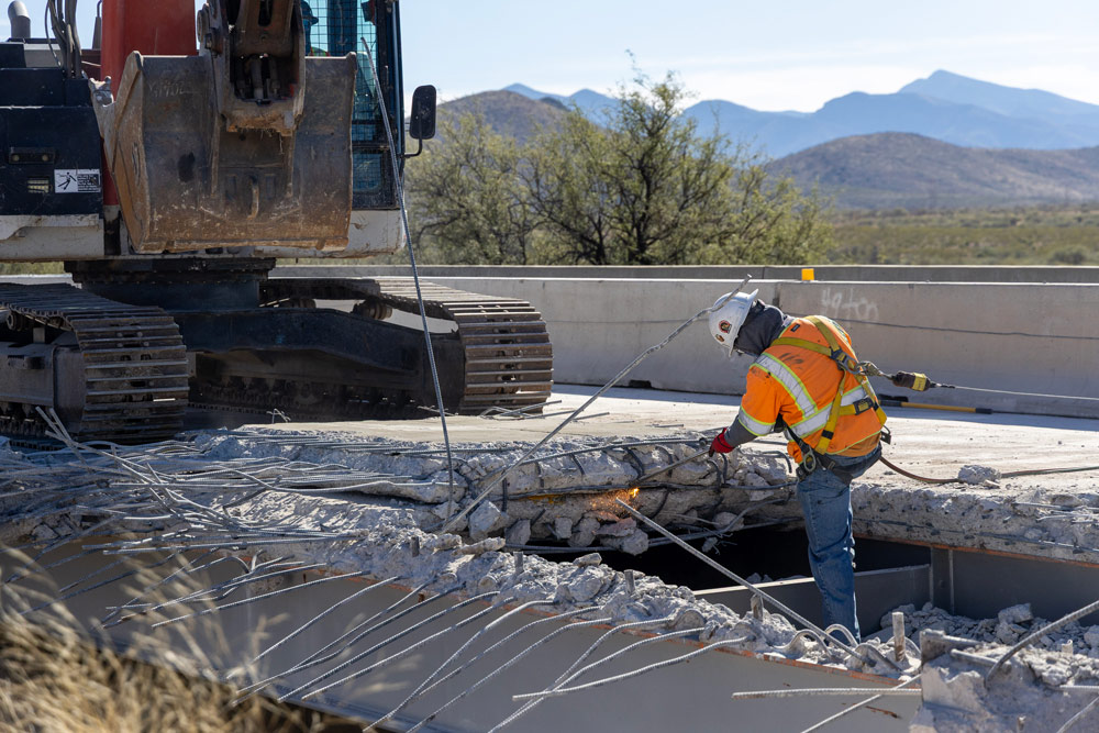 Worker in high-visibility safety gear cutting exposed rebar on a partially demolished bridge deck during the I-10: Amole TI and UPRR Bridge Rehabilitation Project.