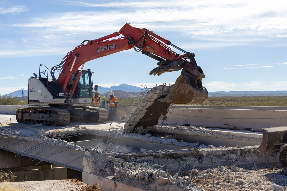 Excavator lifting broken concrete panels from the bridge deck while crew members stand nearby during the I-10: Amole TI and UPRR Bridge Rehabilitation Project.